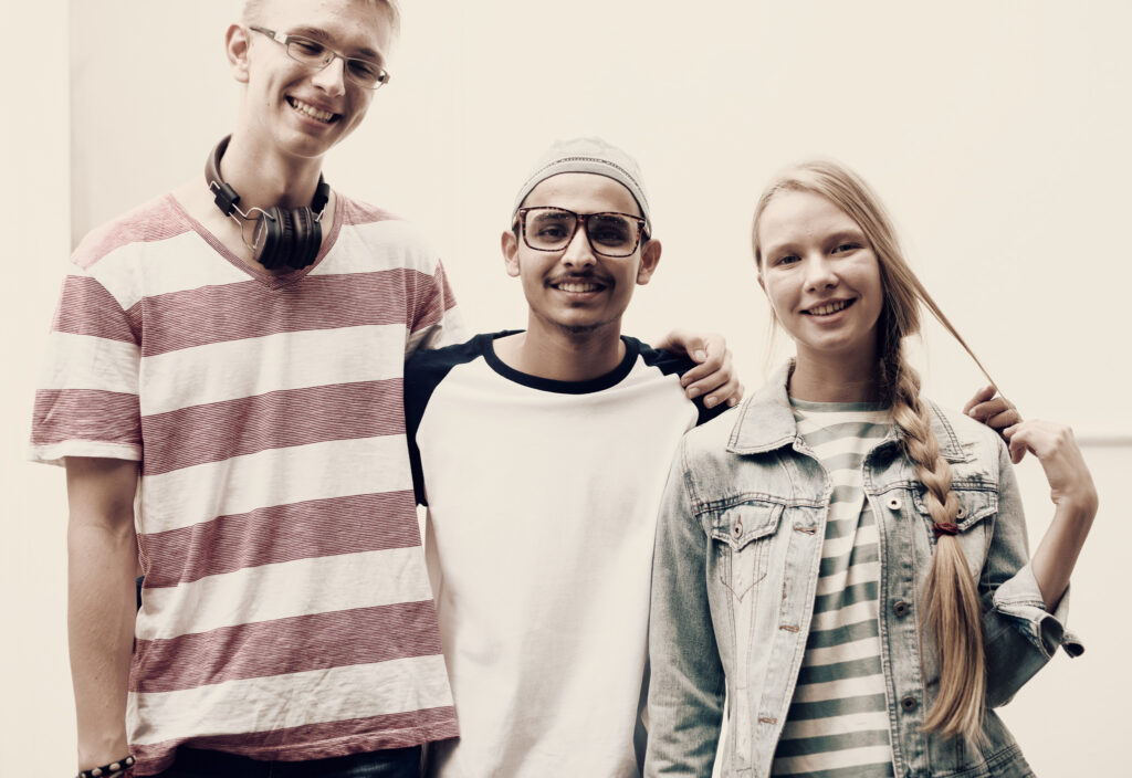 Image of three young adults smiling. They are of diverse nationalities - on the left is a young person with short blonde hair and white skin smiling, wearing glasses, headphone around their neck.