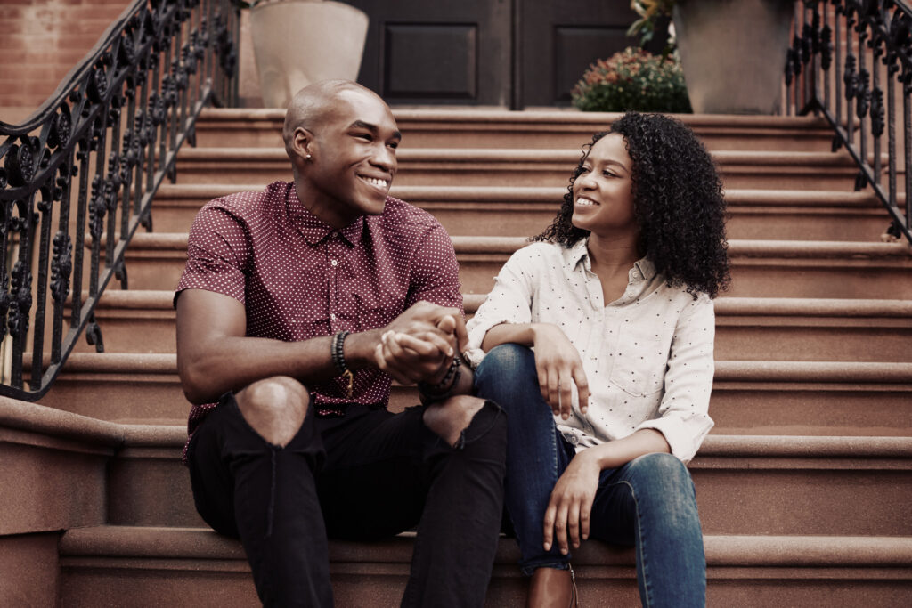 Image of couple sitting on stoop of brownstone in New York city. The man is bald with brown skin, wearing a button up maroon and white dotted button up shirt and black torn denims. The woman has brown curly hair with brown skin, wearing and cream and black dotted button up shirt and blue denims.
