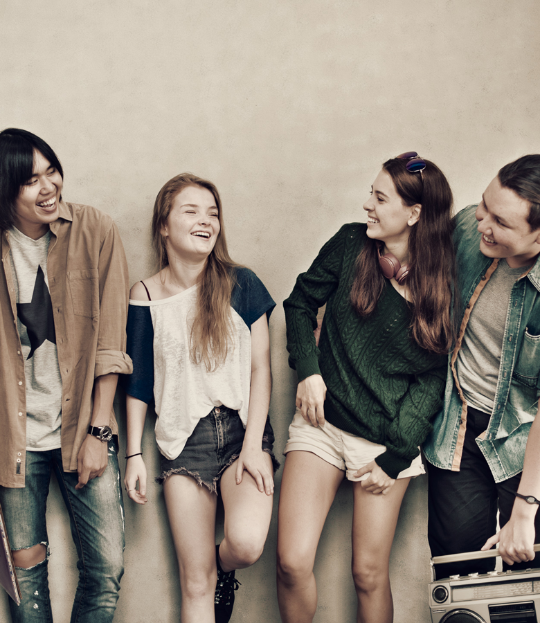 Image of four teenagers leaning against a wall laughing. The teenagers are of diverse cultures and genders.