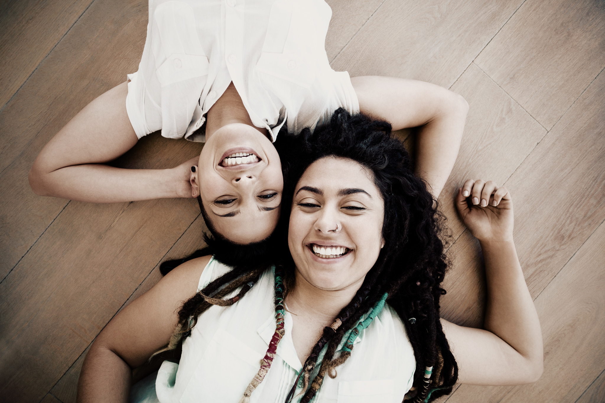Image of two ladies lying on the floor, head to head, looking up at the camera. Both are wearing white shirts.